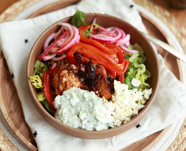 A bowl of salad with grilled chicken, sliced red peppers, pickled onions, crumbled feta cheese, and a serving of tzatziki sauce on a cloth napkin.