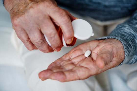An elderly man puts a singular white tablet into the palm of his hand. Only his hands are showing in the photo.