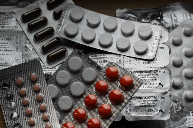 A stack of filled pill packages showing an array of medication sit on the table. The pills range in sizes and colours, from capsules to coated and non-coated. 