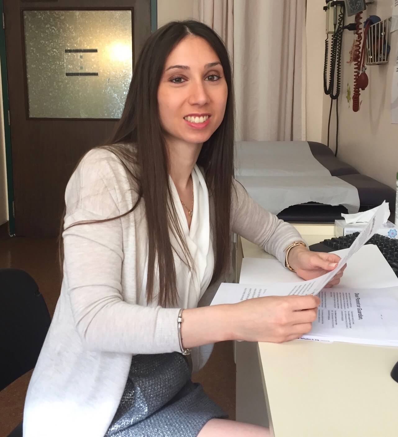 Amanda sits at a desk in a hospital room, going over some papers. She is smiling at the camera. Behind her is an examination bed with paper draped over it. In the background are a variety of medical tools .